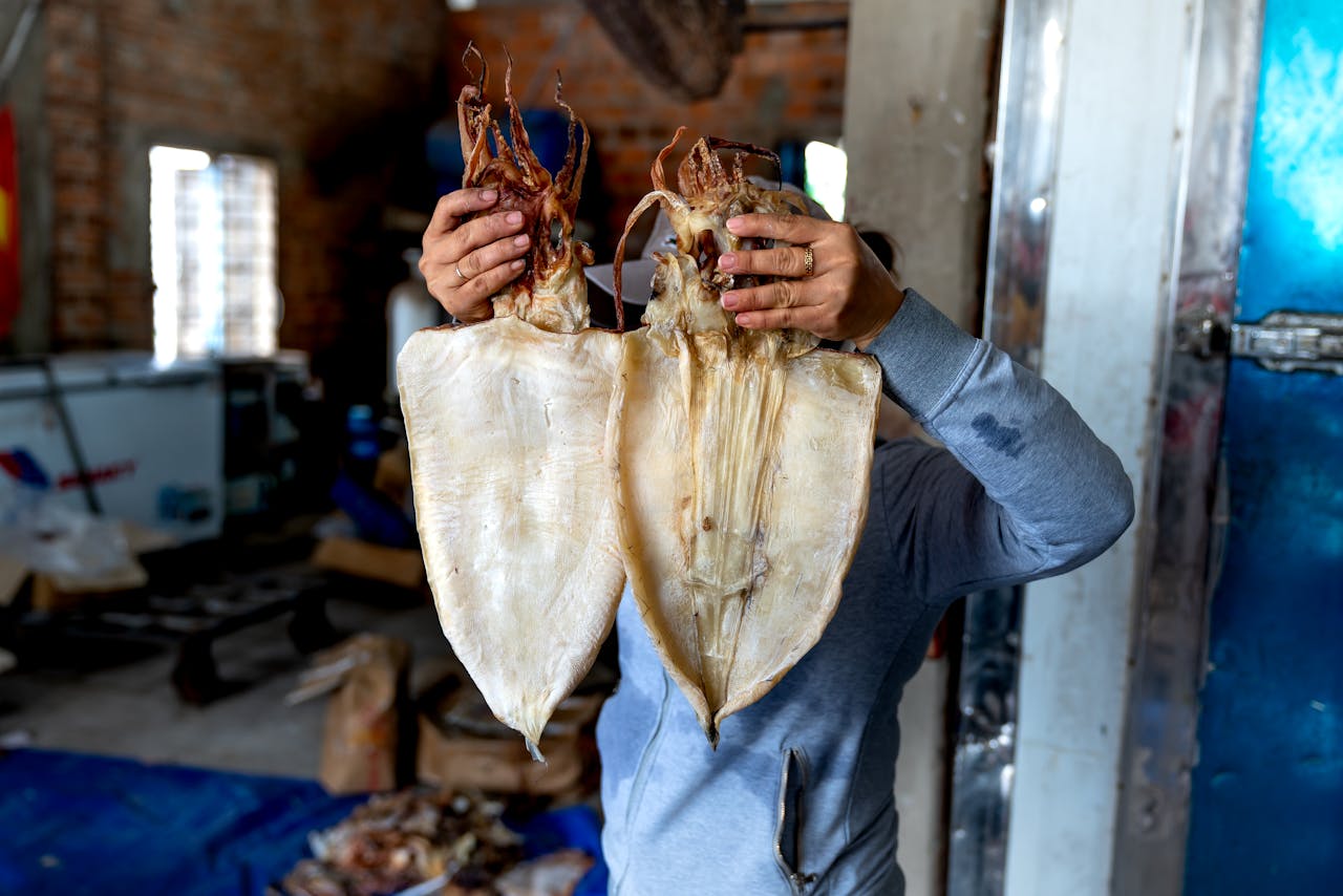 A person holds two large dried squids in an indoor warehouse environment, showcasing seafood preparation.